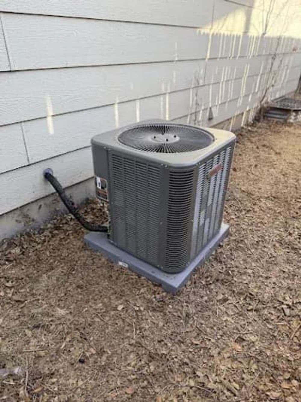 Air conditioner unit installed beside a house on gravel ground, with visible hose connection.